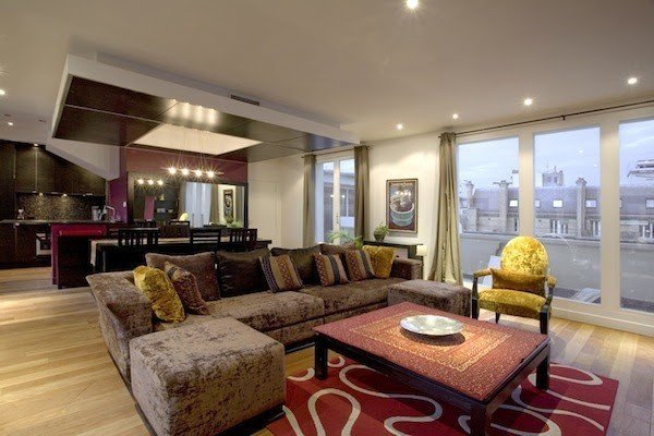 A wide view of the salon with a corner sofa, patterned rug, and open-plan kitchen beyond.